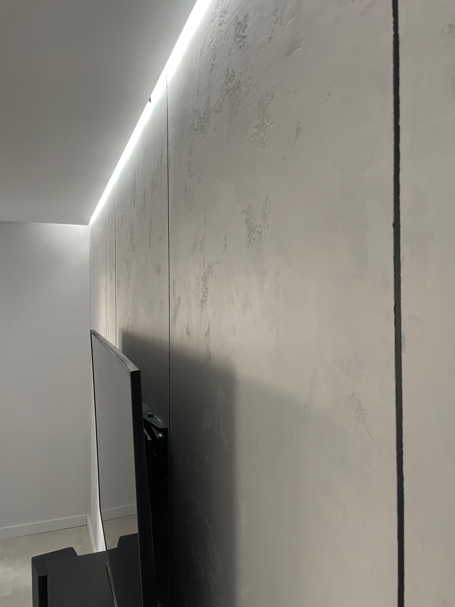 Ściana z dekoracyjną fakturą w odcieniach szarości, oświetlona listwą LED, z widocznym fragmentem telewizora na ścianie.