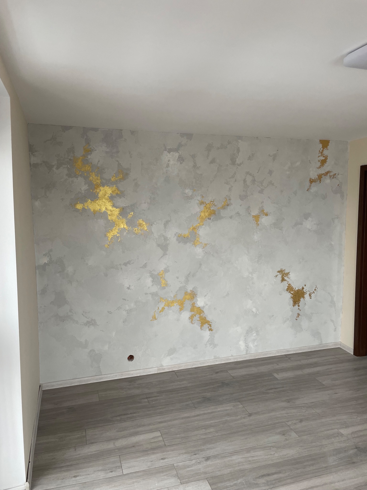Ściana w pokoju z szarą, nieregularną fakturą przypominającą chmury, wzbogacona o złote akcenty, tworzące artystyczny wzór. Podłoga wyłożona szarymi panelami imitującymi drewno.