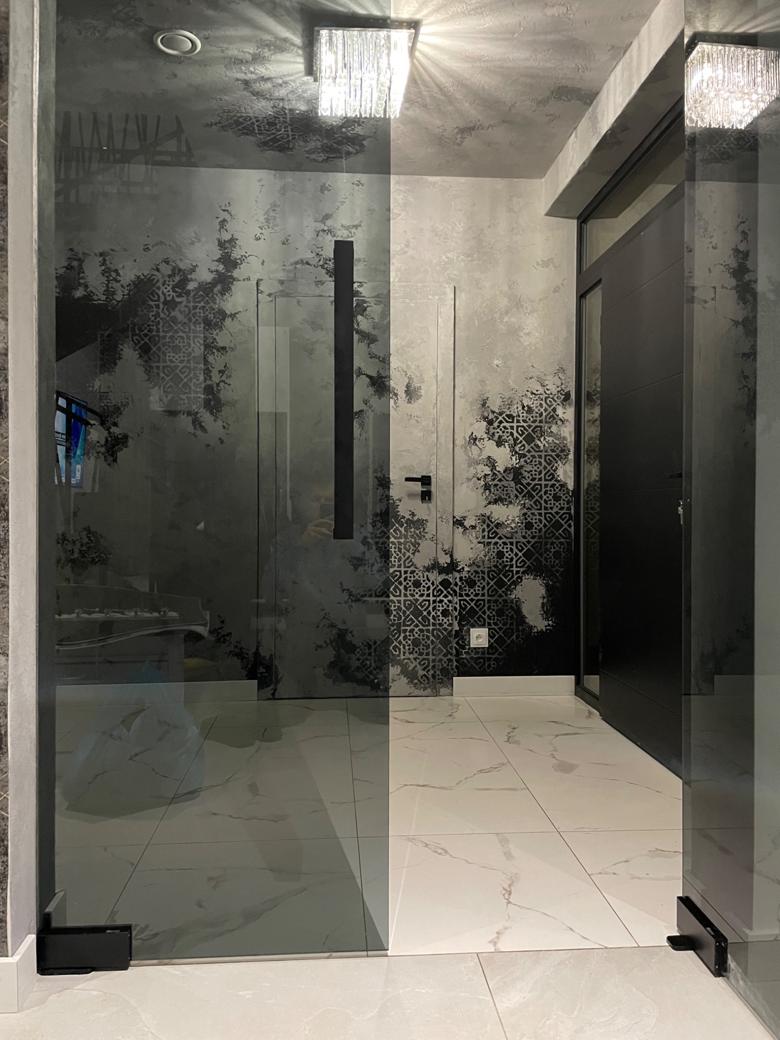 Nowoczesne wnętrze z dekoracyjną ścianą w odcieniach szarości, wzór przypominający marmur z geometrycznymi elementami, szklane drzwi z czarnym uchwytem, biała podłoga z marmurowym wzorem, widoczne...