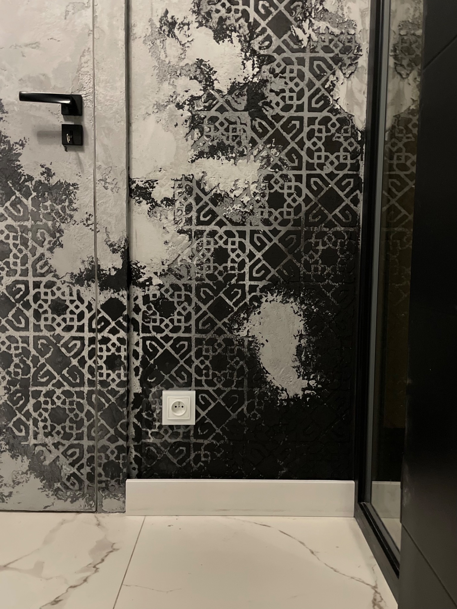 Ściana z dekoracyjną fakturą w odcieniach szarości i czerni, wzór geometryczny, widoczny biały kontakt elektryczny i fragment białej listwy przypodłogowej na tle marmurowej podłogi.