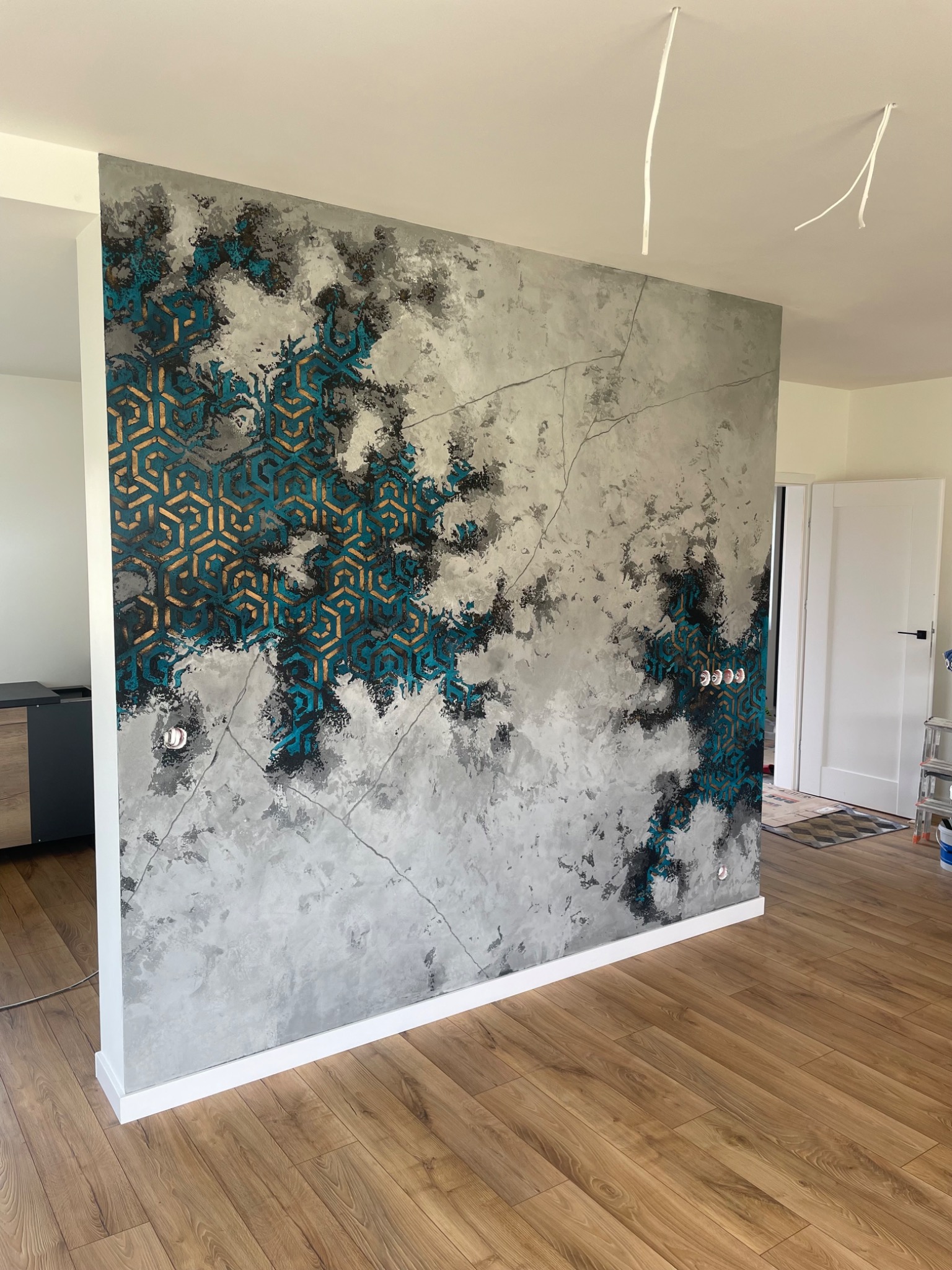 Ściana działowa z dekoracyjnym tynkiem w odcieniach szarości, czerni i turkusu z geometrycznym wzorem, widoczne gniazdka elektryczne i fragment drewnianej podłogi.