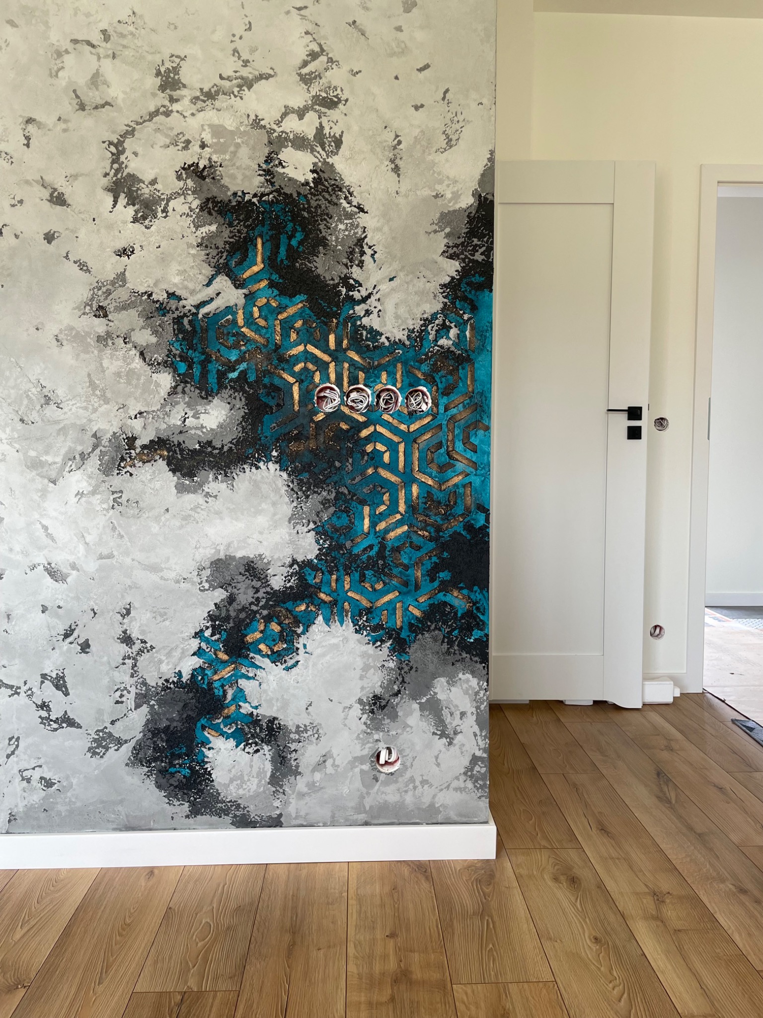 Ściana z dekoracyjną fakturą przypominającą chmury w odcieniach szarości i błękitu, z geometrycznym wzorem w kolorze złotym, na tle białych drzwi i drewnianej podłogi.