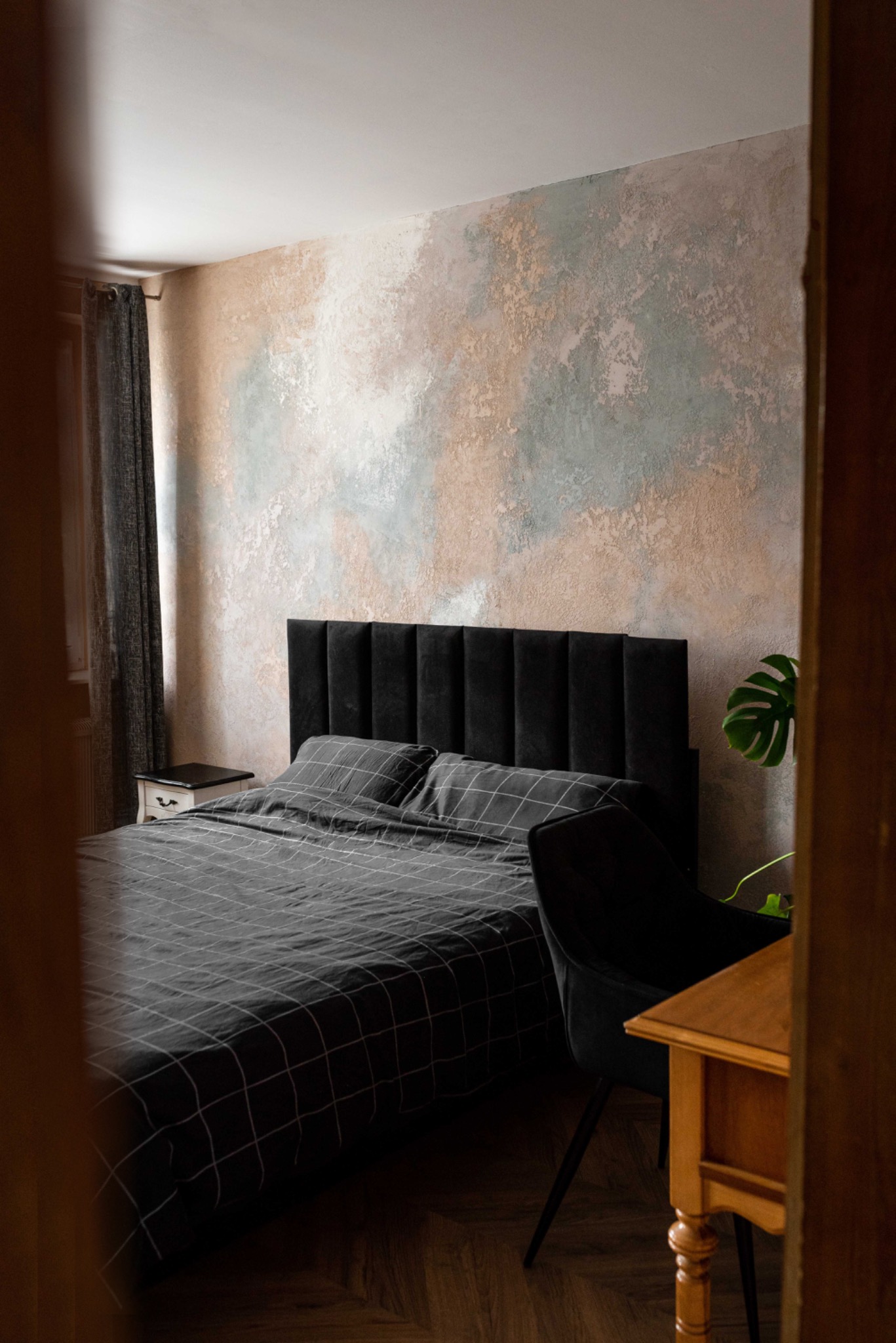Sypialnia z łóżkiem z czarnym wezgłowiem i pościelą w kratę, ściana dekoracyjna z efektem malarskim w odcieniach beżu i szarości, widok zza framugi drzwi.