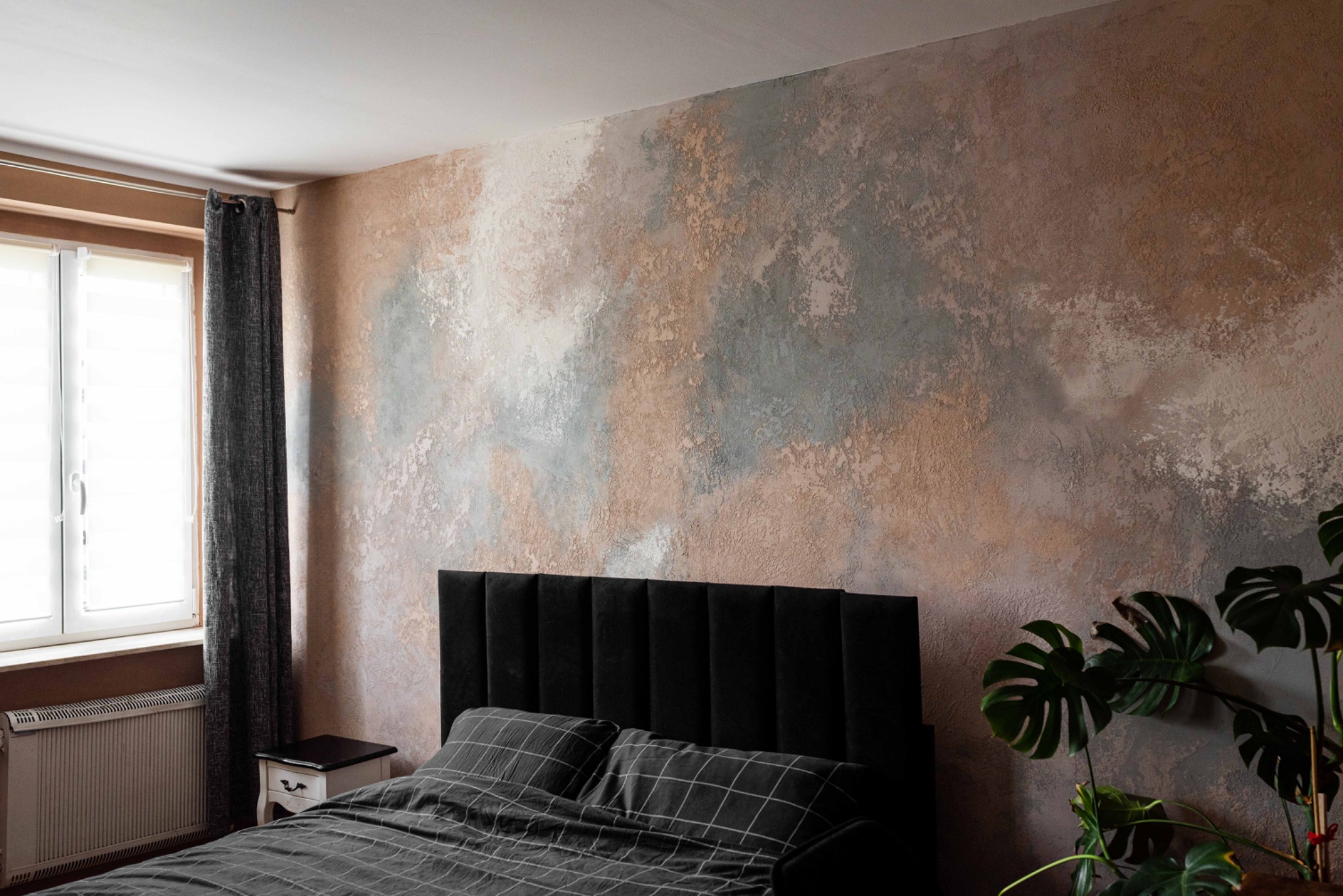 Ściana w sypialni z dekoracyjną fakturą w odcieniach beżu, szarości i bieli, imitująca naturalny kamień lub chmurę, widoczna nad czarnym, tapicerowanym zagłówkiem łóżka i rośliną Monstera.