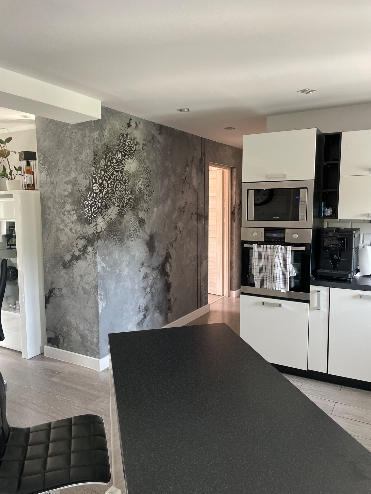 Wykończona ściana w kuchni z dekoracyjną fakturą w odcieniach szarości i srebrnym ornamentem, widok na nowoczesną zabudowę kuchenną z białymi szafkami i sprzętem AGD.
