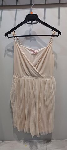 Kremowa sukienka na ramiączkach z plisowanym dołem, wisząca na czarnym wieszaku na tle szarej ściany.