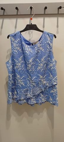 Jasnoniebieska bluzka damska bez rękawów z białym haftem kwiatowym, wisząca na czarnym wieszaku na tle szarej ściany.