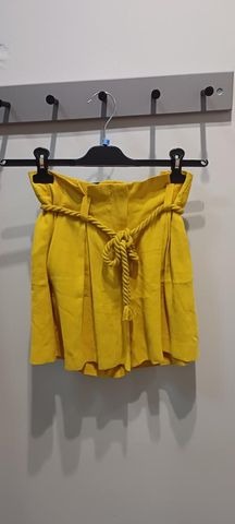 Żółte, plisowane szorty z wiązanym sznurkowym paskiem, wiszące na czarnym wieszaku na tle jasnoszarej ściany z haczykami.