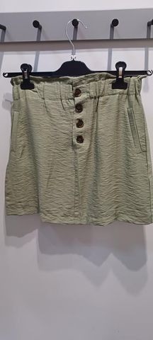 Krótka, pistacjowa spódnica z wysokim stanem i guzikami, wisząca na wieszaku na tle jasnej ściany.