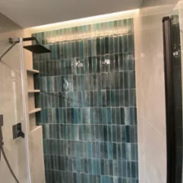 Wykończona kabina prysznicowa z turkusową, pionową glazurą i podświetleniem LED. Widoczna czarna deszczownica i szklane drzwi. Czyste, nowoczesne wykończenie łazienki.