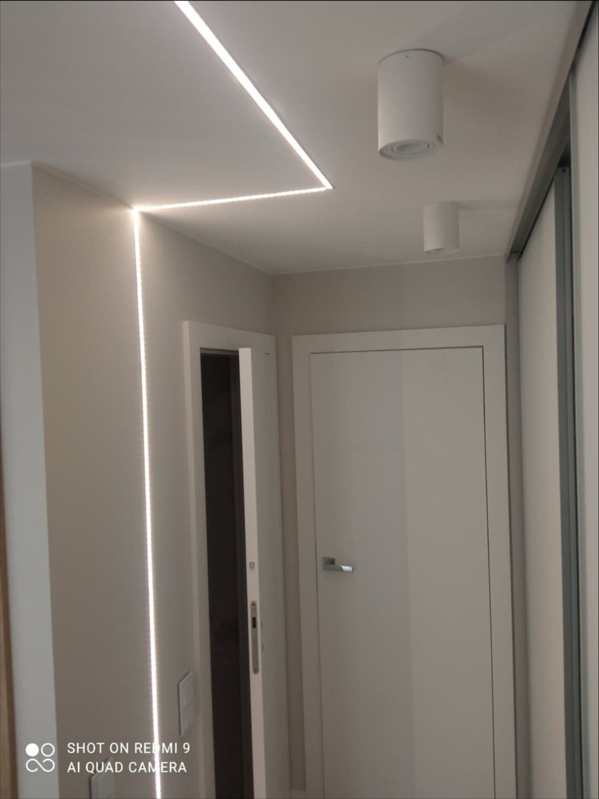 Nowoczesny korytarz z oświetleniem LED wpuszczonym w sufit, punktowe lampy sufitowe, białe drzwi wewnętrzne i przesuwne drzwi do szafy.