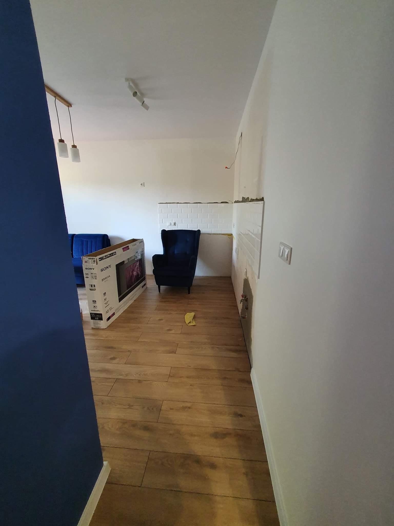 Wnętrze w trakcie prac wykończeniowych: położona drewnopodobna podłoga, częściowo wykończona ściana z białymi płytkami, widoczna puszka elektryczna, karton po telewizorze Sony Bravia, niebieski...