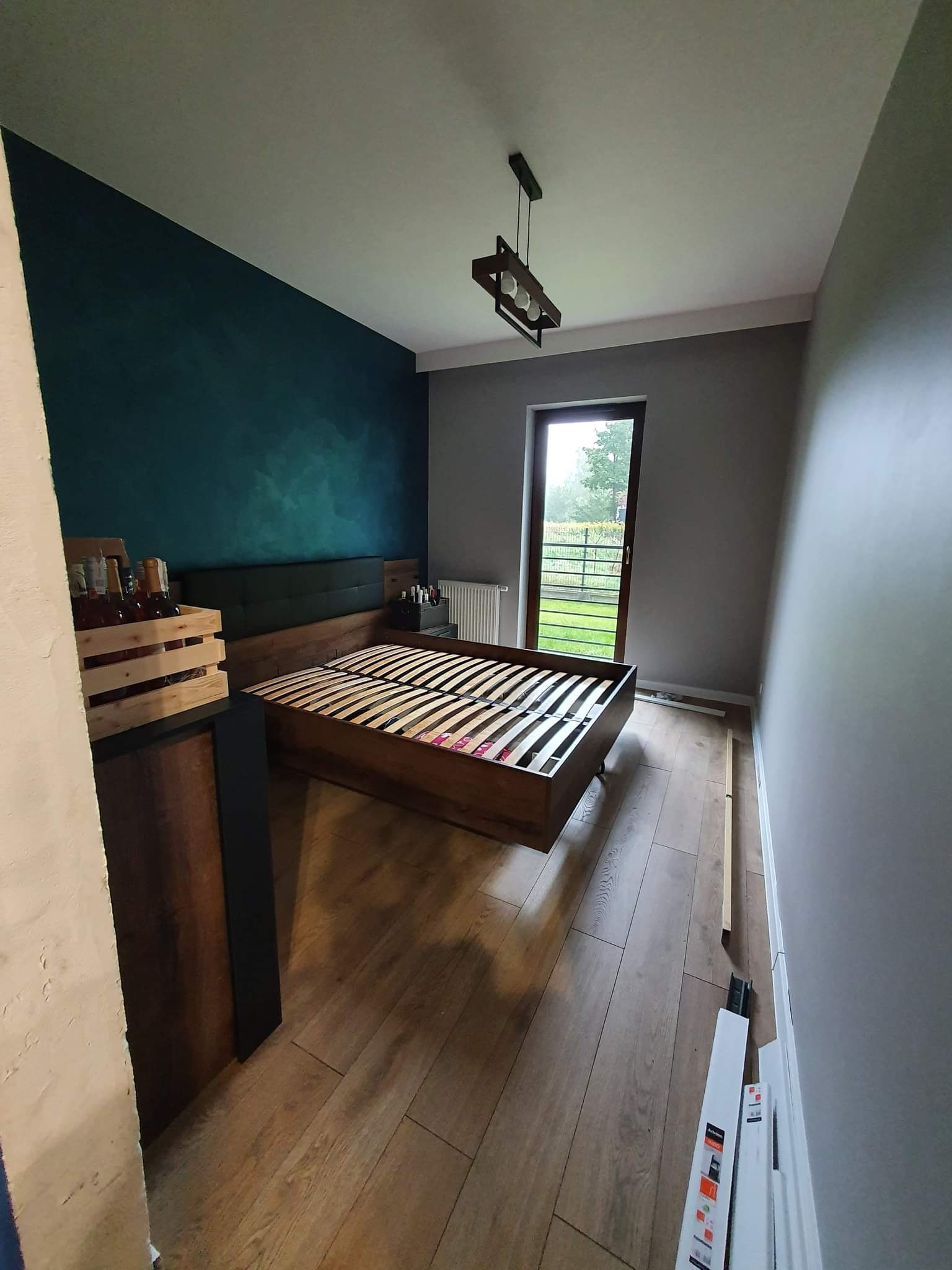 Sypialnia w trakcie urządzania: drewniany stelaż łóżka bez materaca na tle ściany w kolorze butelkowej zieleni i szarej ściany, widoczne kartony przy drzwiach balkonowych, lampa sufitowa w stylu...