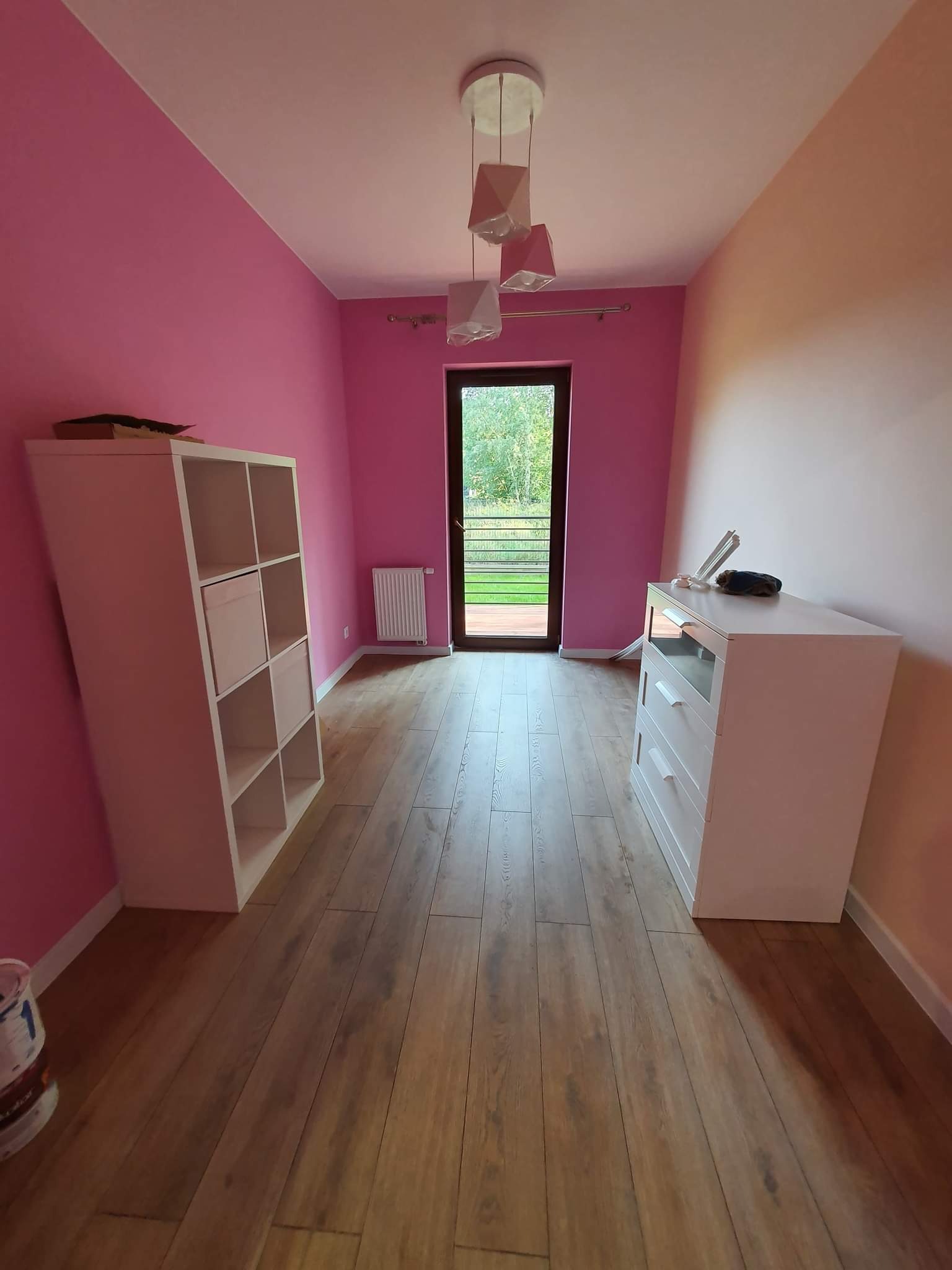 Pokój po odświeżeniu, ściany pomalowane na różowo i brzoskwiniowo, drewniana podłoga, białe meble i lampa z geometrycznymi kloszami.