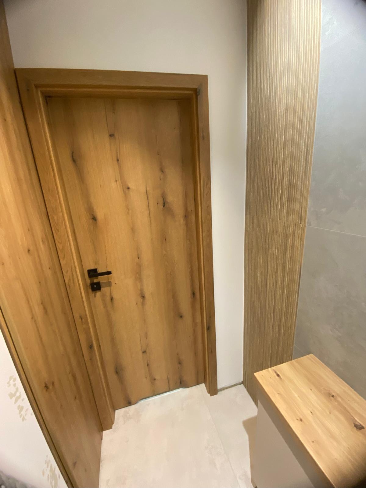 Zamknięte, drewniane drzwi z czarną klamką, dopasowane do drewnianej okładziny ściennej w łazience. Widoczny fragment blatu.