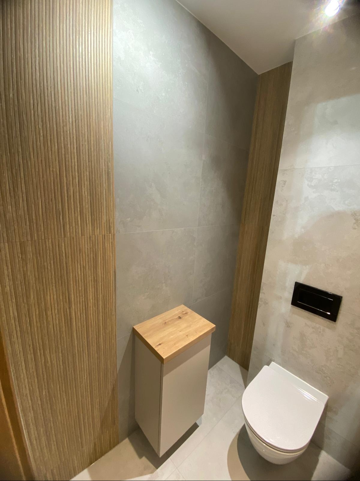 Wykończona łazienka z beżową szafką pod umywalką z drewnianym blatem i toaletą z czarnym przyciskiem spłukującym, na ścianach szare płytki i dekoracyjne panele.