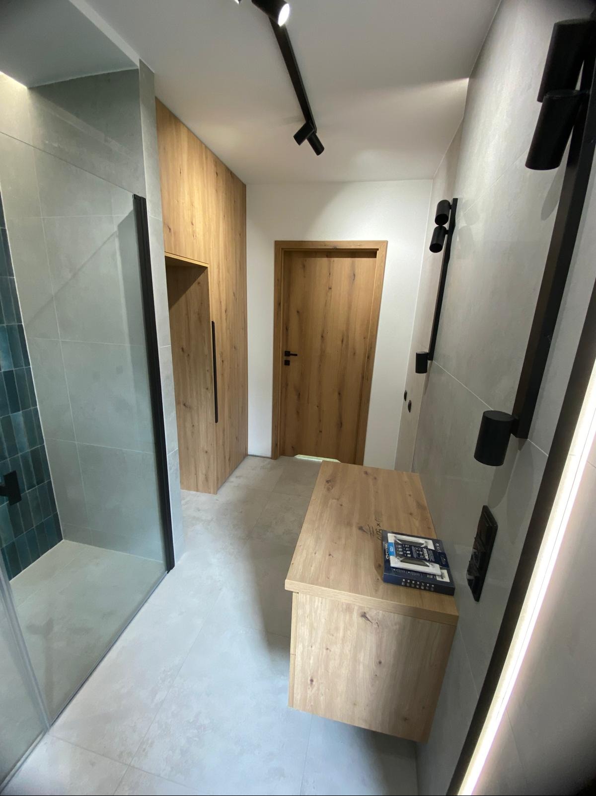 Nowoczesna łazienka po remoncie: kabina prysznicowa z ciemnozieloną mozaiką, szafka z blatem z imitacji drewna i czarne oświetlenie punktowe.