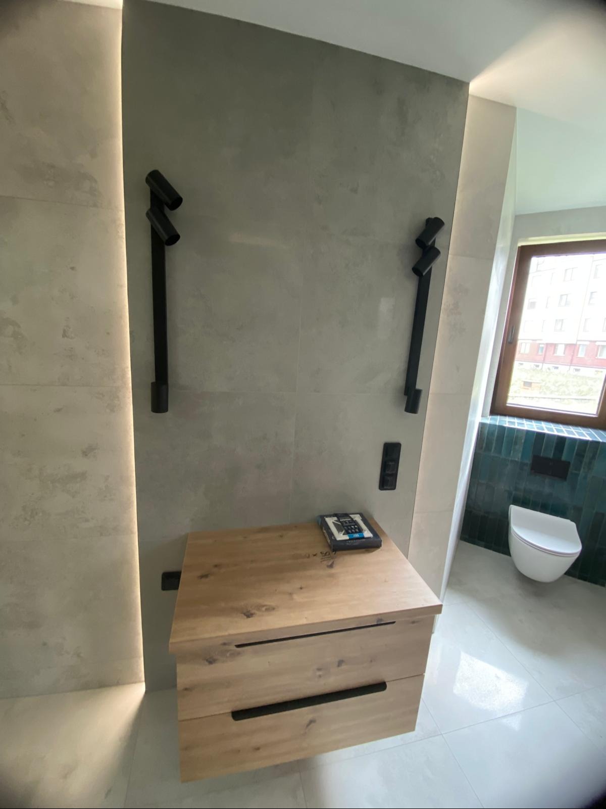 Nowoczesna łazienka po remoncie: drewniana szafka z czarnymi uchwytami, szare płytki, czarne lampy ścienne i toaleta w tle. Widok na okno.