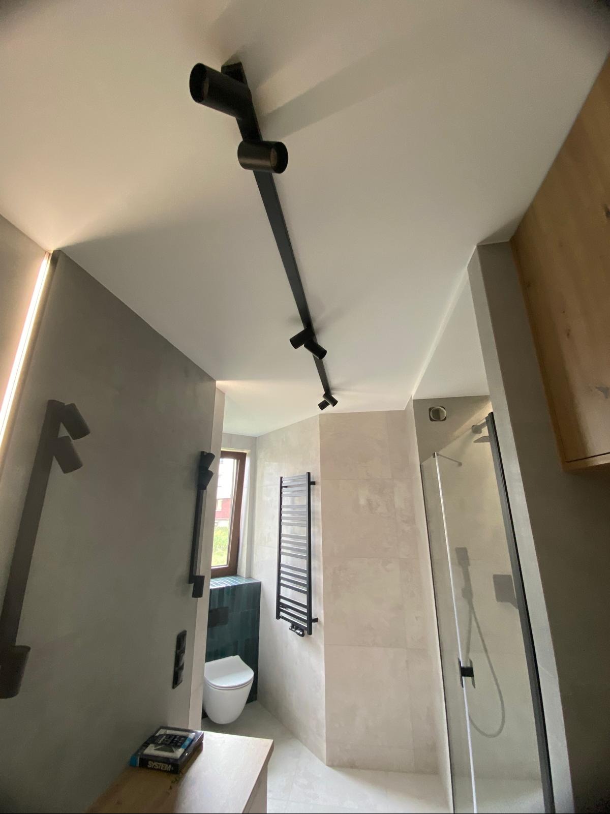 Nowoczesna łazienka z czarnym oświetleniem szynowym, beżowymi płytkami i grzejnikiem drabinkowym. Widoczna kabina prysznicowa i toaleta.