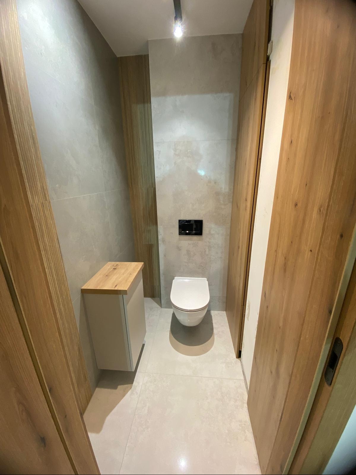 Nowoczesna łazienka po remoncie: wisząca toaleta, szafka z drewnianym blatem, płytki w odcieniach szarości i drewniane drzwi przesuwne.