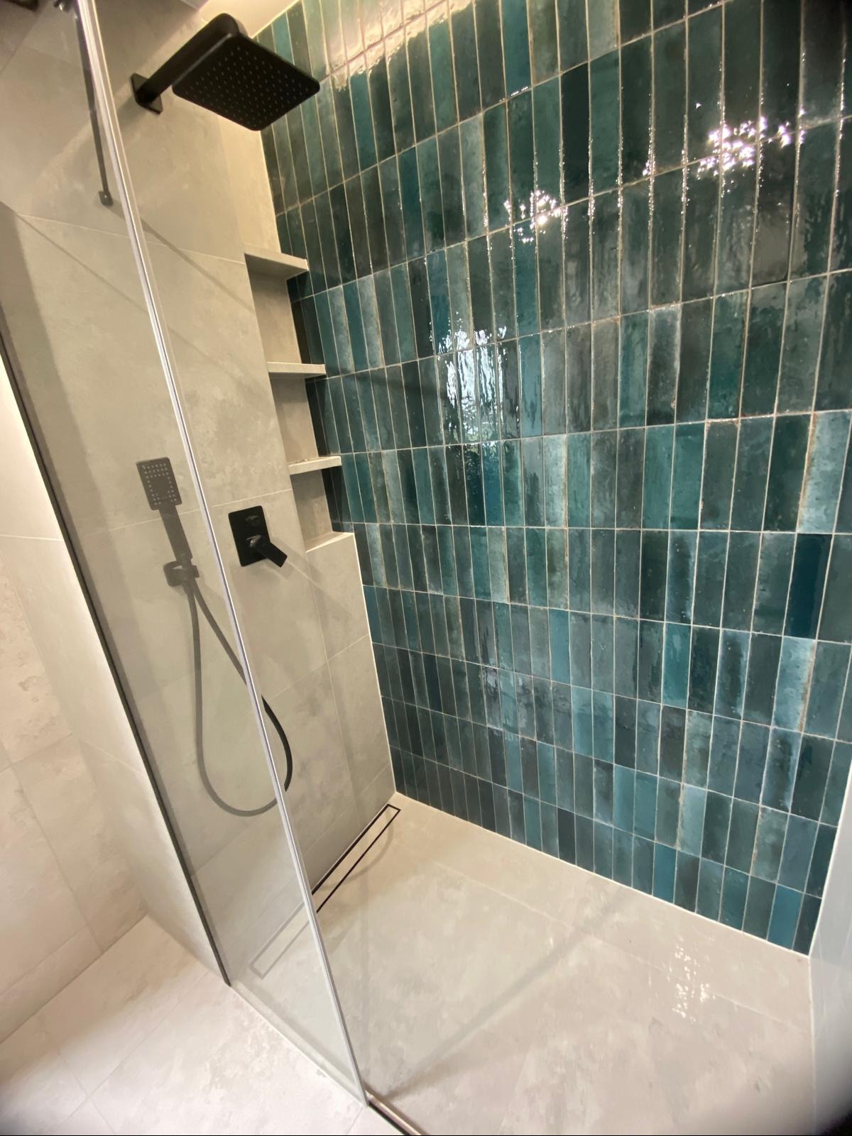 Nowoczesna kabina prysznicowa z turkusowymi, błyszczącymi płytkami ułożonymi pionowo, czarną armaturą i szklaną ścianką. W tle wnęki w ścianie.