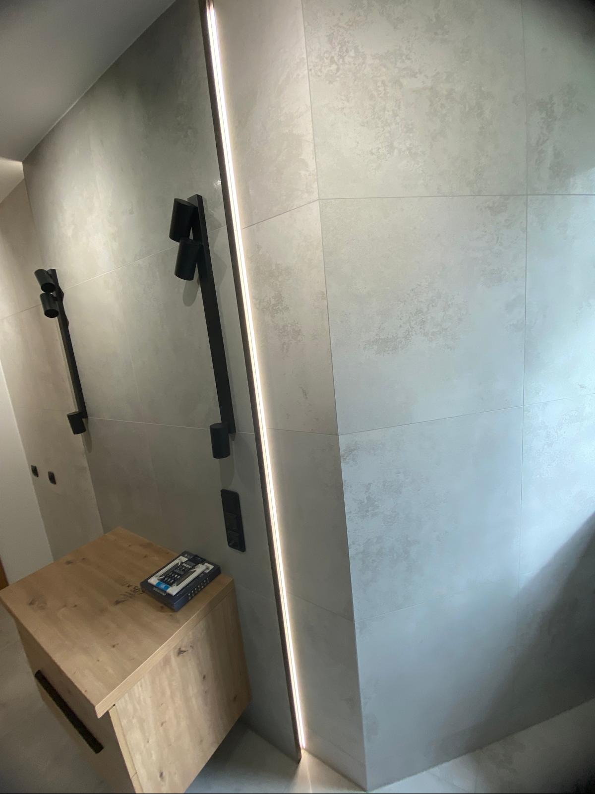 Nowoczesna łazienka z szarymi płytkami, oświetleniem LED wzdłuż ściany i drewnianą szafką z czarnymi uchwytami. Minimalistyczne czarne lampy ścienne.