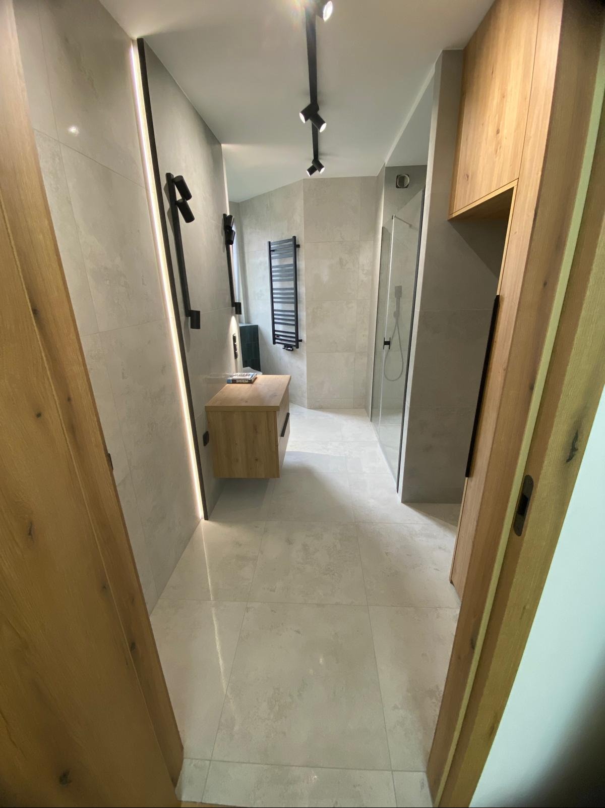 Nowoczesna łazienka z kabiną prysznicową, drewnianą szafką pod umywalką i grzejnikiem drabinkowym, oświetlona punktowymi lampami.