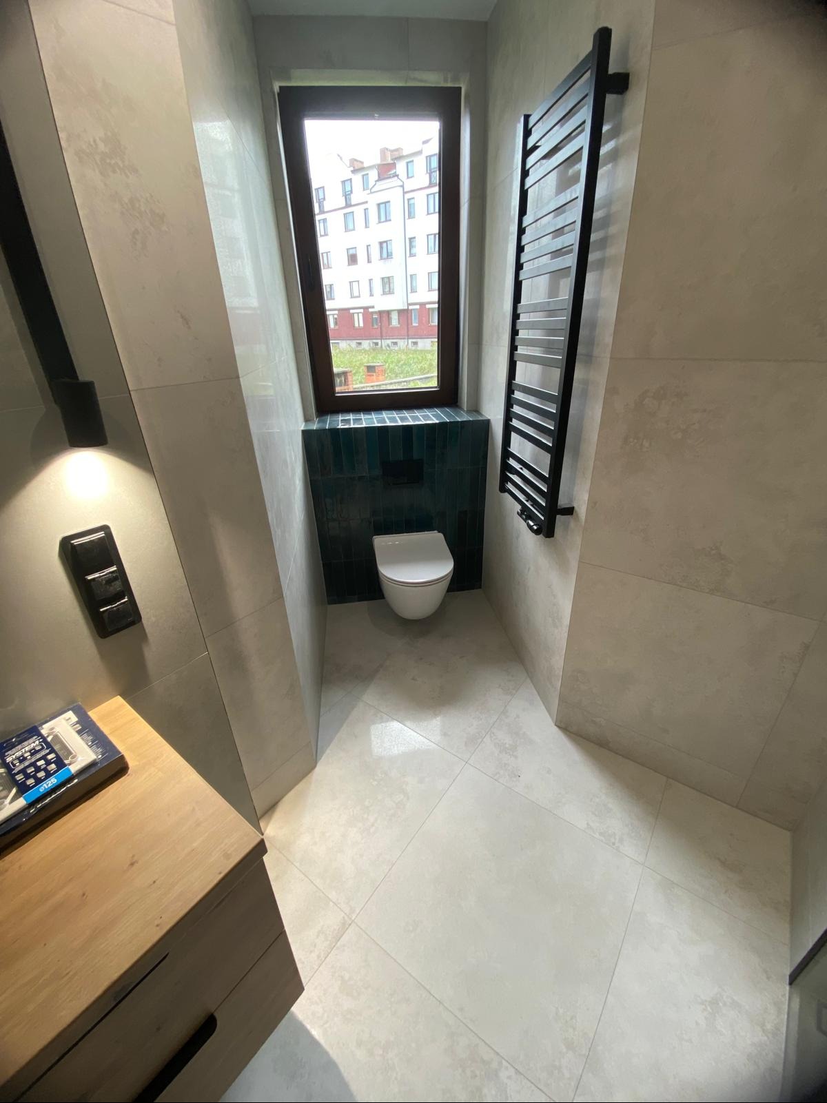 Nowoczesna toaleta w beżowej tonacji z akcentem turkusowych płytek pod oknem. Na ścianie grzejnik drabinkowy w czarnym macie. Widok z góry na wykończone wnętrze.