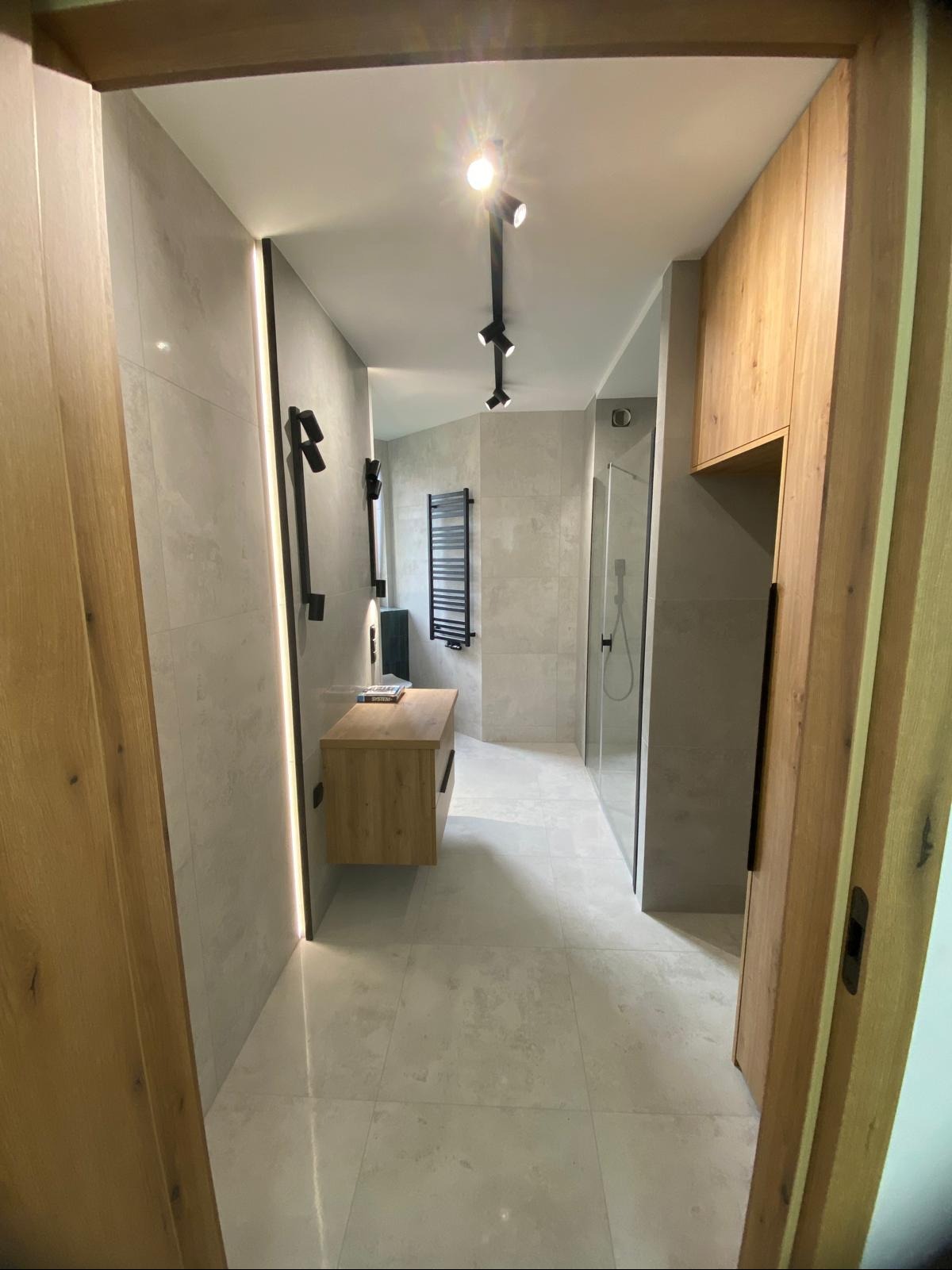 Wykończona łazienka w jasnych barwach. Widoczna drewniana szafka pod umywalką, grzejnik drabinkowy, kabina prysznicowa i oświetlenie punktowe. Nowoczesny design.