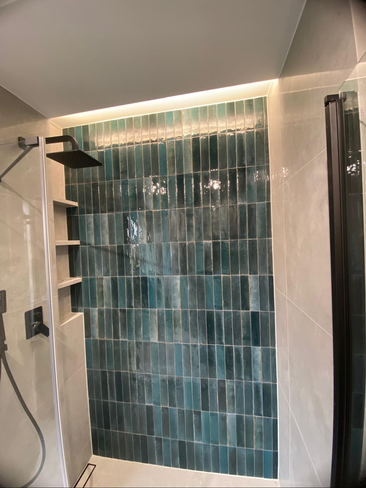 Wykończona kabina prysznicowa z turkusową, pionową glazurą i podświetleniem LED. Widoczna czarna deszczownica i szklane drzwi. Czyste, nowoczesne wykończenie łazienki.