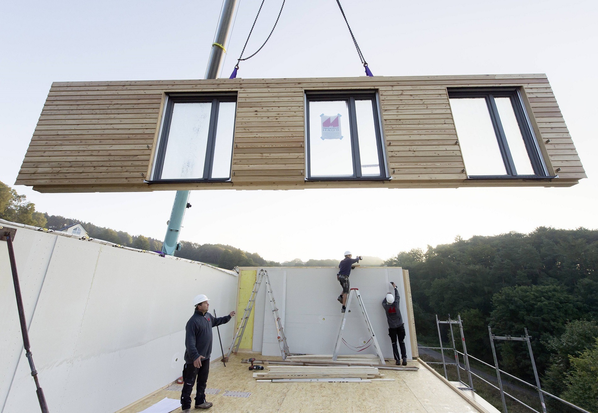 Montaż segmentu domu z drewnianą elewacją i trzema oknami, podnoszonego przez dźwig, pracownicy na drabinach mocują go do ściany budynku.