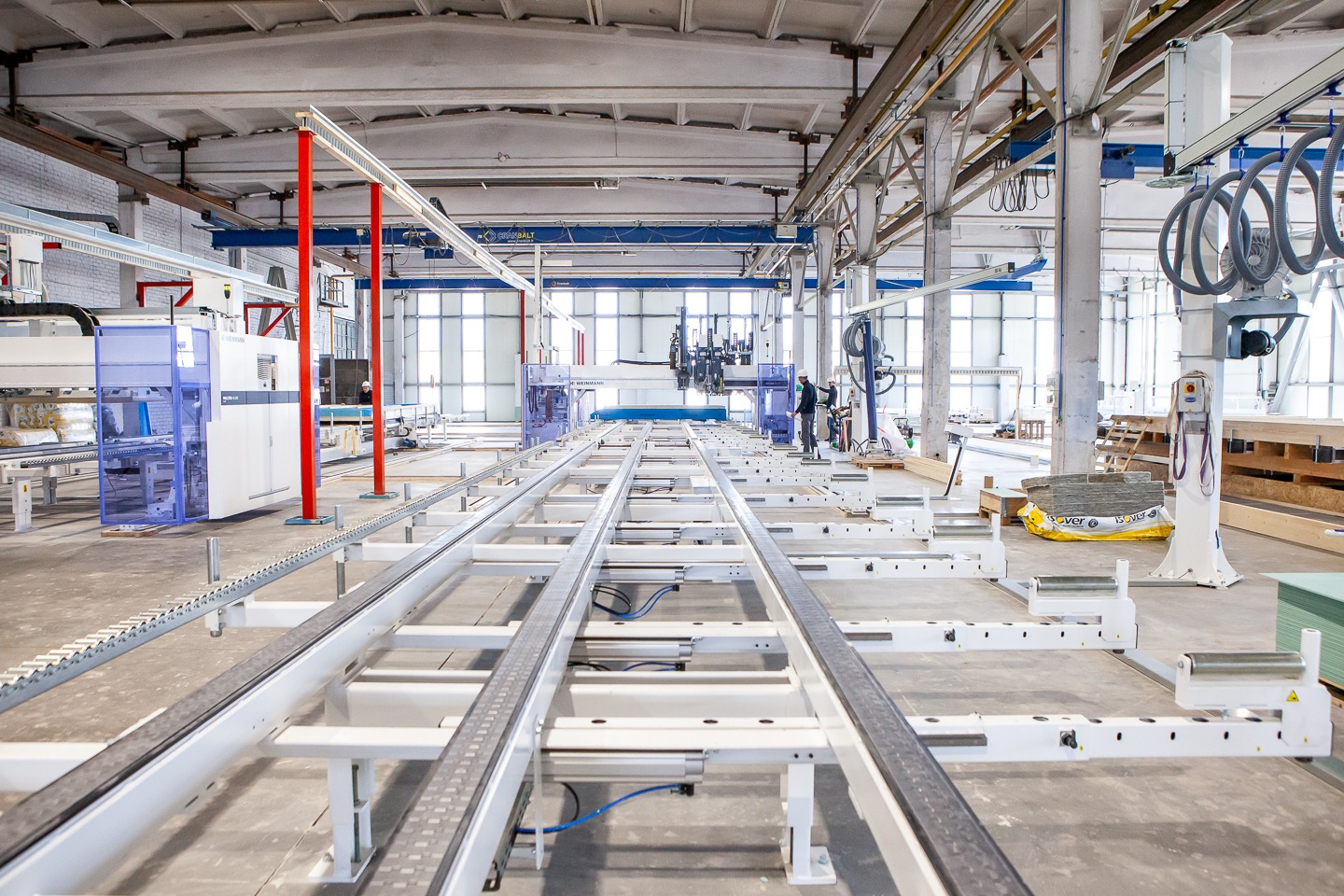 Wnętrze hali produkcyjnej z linią technologiczną do obróbki elementów drewnianych, widoczne maszyny CNC i pracownicy w tle, oświetlenie dzienne przez duże okna.