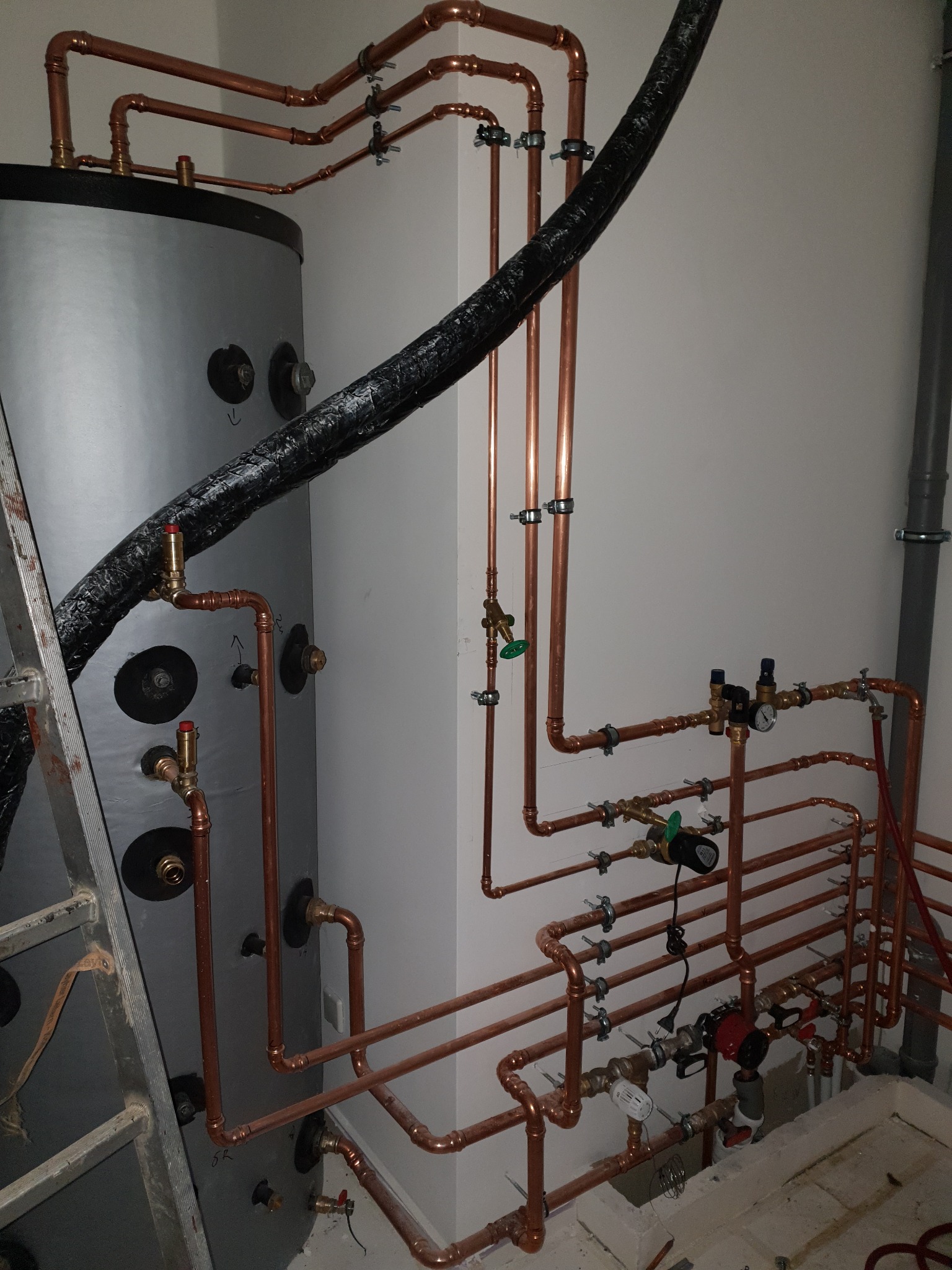 Nowa instalacja grzewcza z miedzianych rur, zaworami i czujnikami, podłączona do szarego zbiornika buforowego, widoczna drabina aluminiowa.