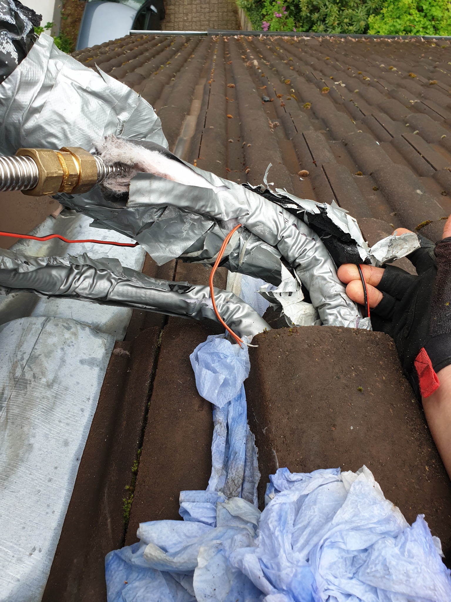 Izolacja rur na dachu pokrytym dachówką, zabezpieczona srebrną taśmą, widoczne kable i fragment dłoni w rękawicy ochronnej.