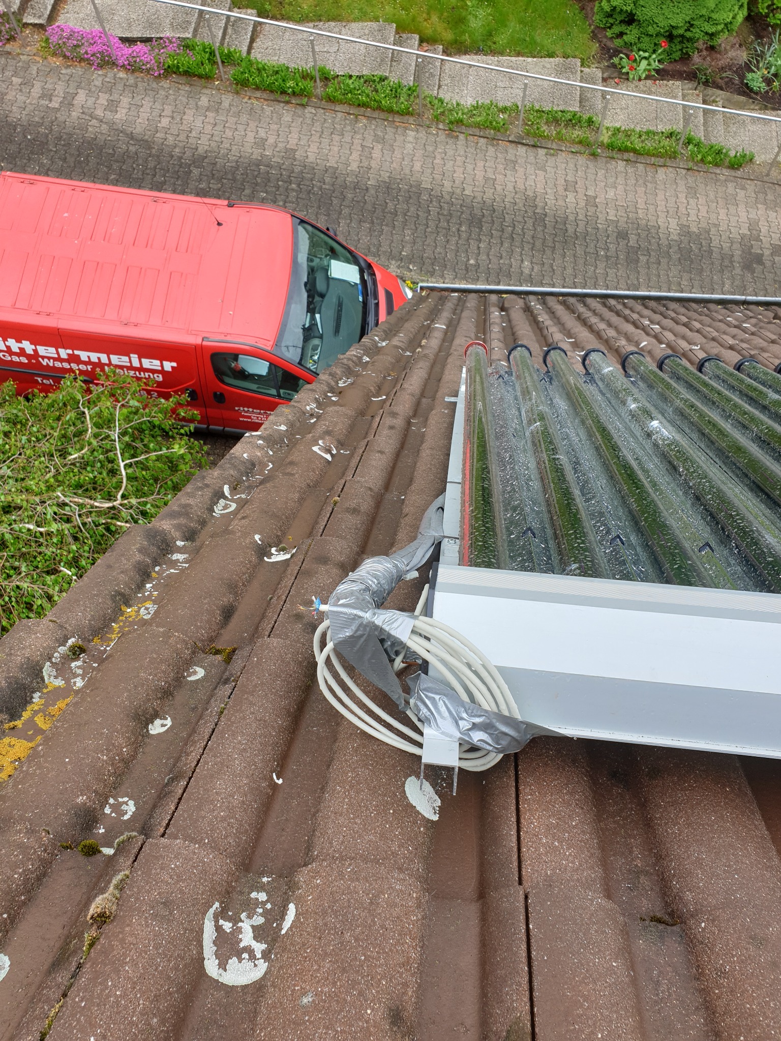 Widok z góry na zamontowane kolektory słoneczne rurowe na brązowym dachu pokrytym dachówką, z widocznym czerwonym samochodem dostawczym firmy instalacyjnej i fragmentem ogrodu.