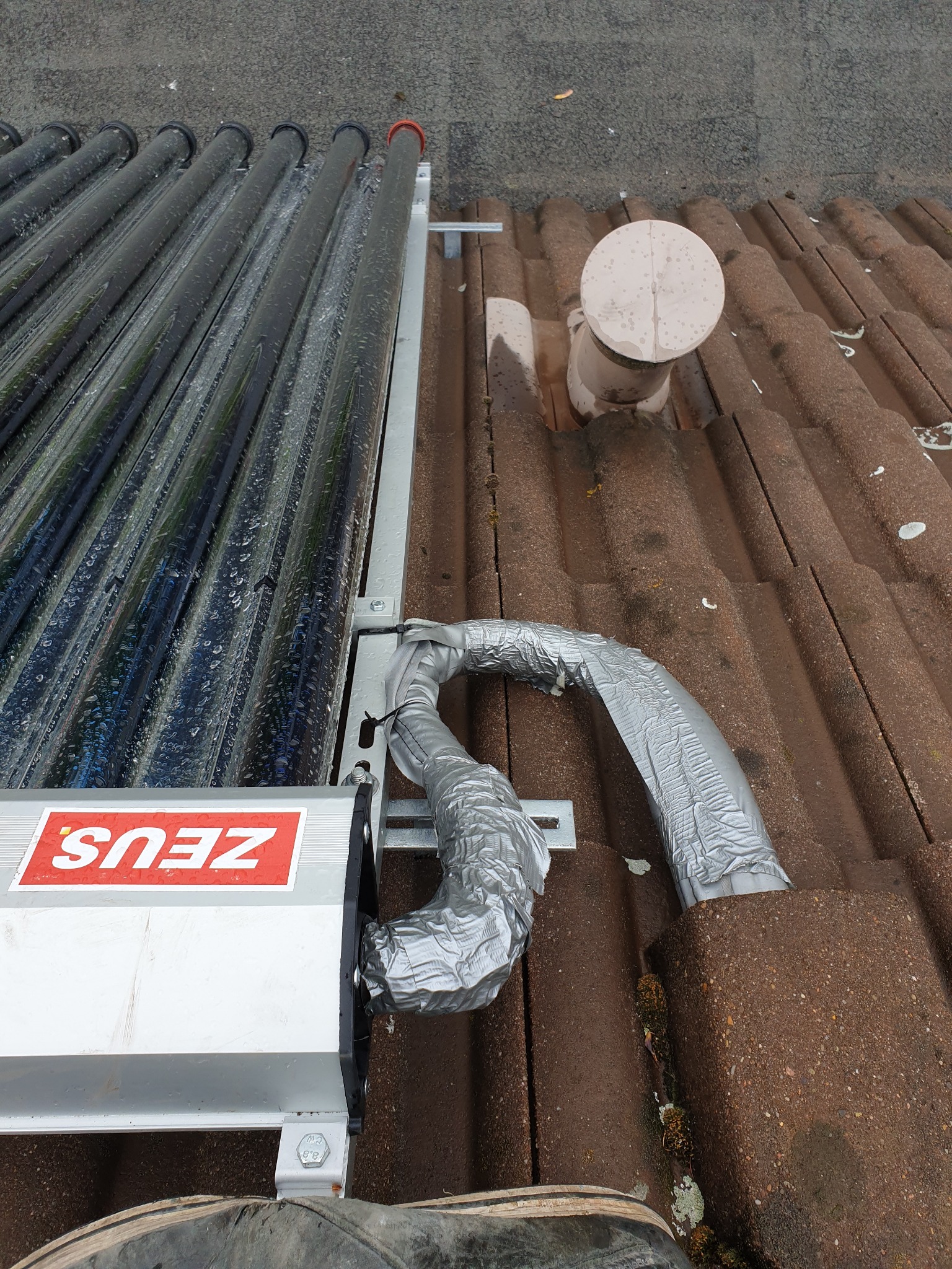 Panel solarny z rurami próżniowymi na brudnym, brązowym dachu pokrytym dachówką, z widocznym kominem wentylacyjnym i izolowanymi przewodami.