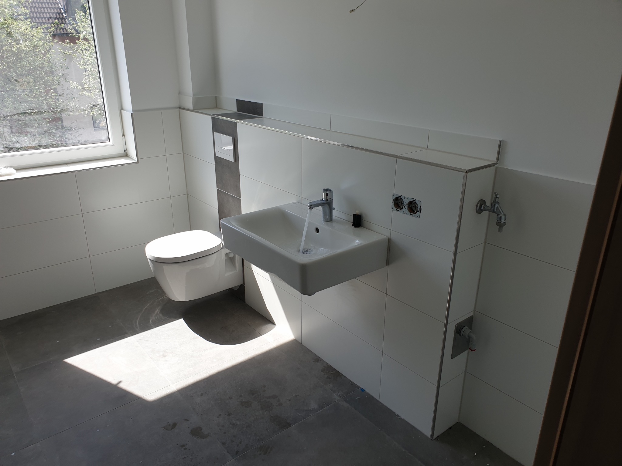 Nowoczesna łazienka w trakcie wykańczania: podwieszana toaleta, umywalka z baterią, wylewająca się woda, płytki ceramiczne na ścianach, szare płytki podłogowe, widoczne gniazdka elektryczne...