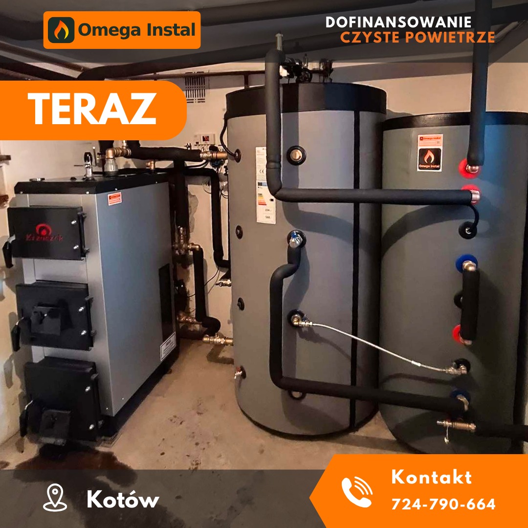 Szary piec na paliwo stałe z trzema drzwiczkami i dwoma zbiornikami akumulacyjnymi w kotłowni, widoczne rury i armatura, na ścianie naklejka informacyjna o dofinansowaniu.