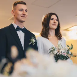 Elegancka para młoda podczas ceremonii: pan młody w granatowym smokingu, panna młoda w białej sukni z bukietem. Delikatne światło, subtelny uśmiech.