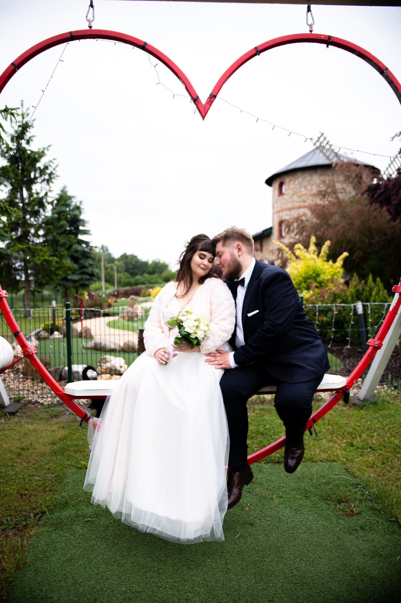 Para młoda siedzi na huśtawce w kształcie serca, w tle widać budynek przypominający wiatrak. Romantyczna sesja ślubna w plenerze.