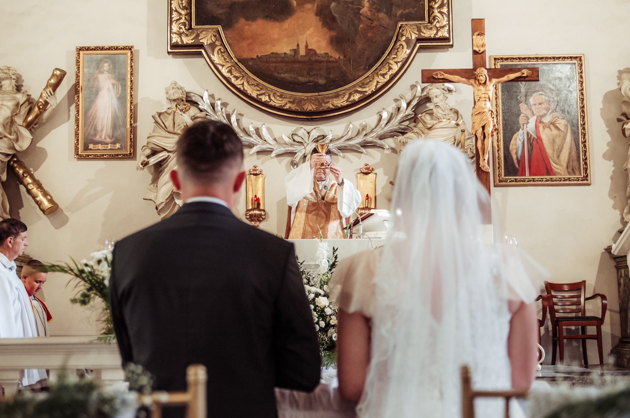 Para młoda w kościele podczas ceremonii ślubnej. Ksiądz w bogato zdobionym ornacie odprawia mszę. W tle obrazy i krucyfiks, widok zza pleców nowożeńców.
