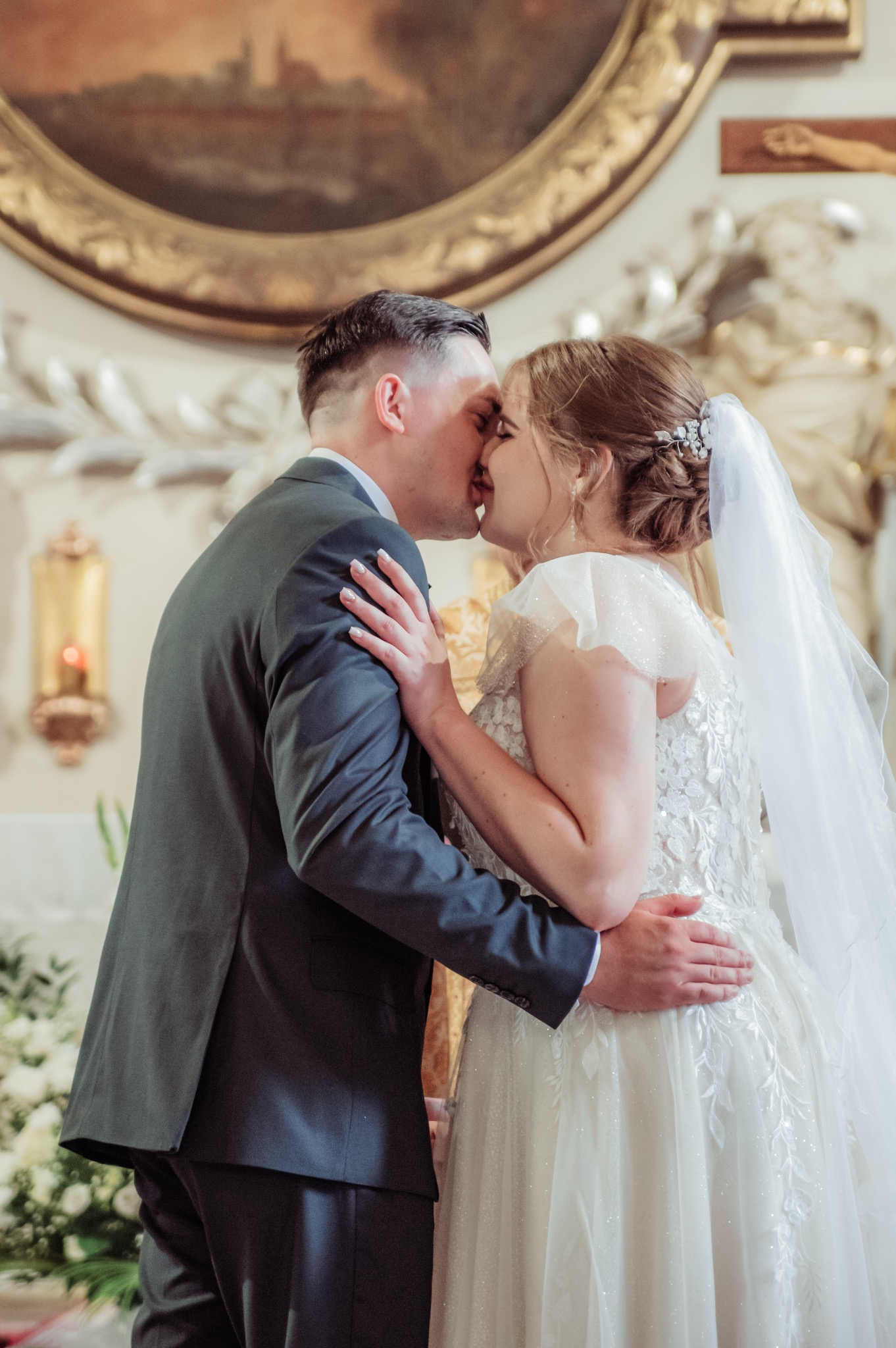 Pocałunek pary młodej w kościele na tle dekoracji i obrazu w złotej ramie. Panna młoda w sukni z welonem, pan młody w garniturze. Romantyczny moment.
