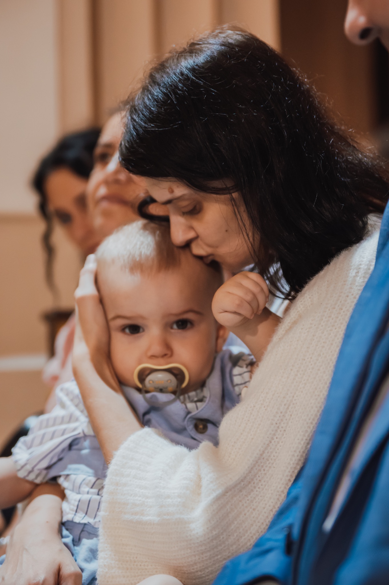 Matka całuje w głowę niemowlę z smoczkiem, obecni goście w tle. Ujęcie z uroczystości rodzinnej, ciepłe światło, delikatne kolory.