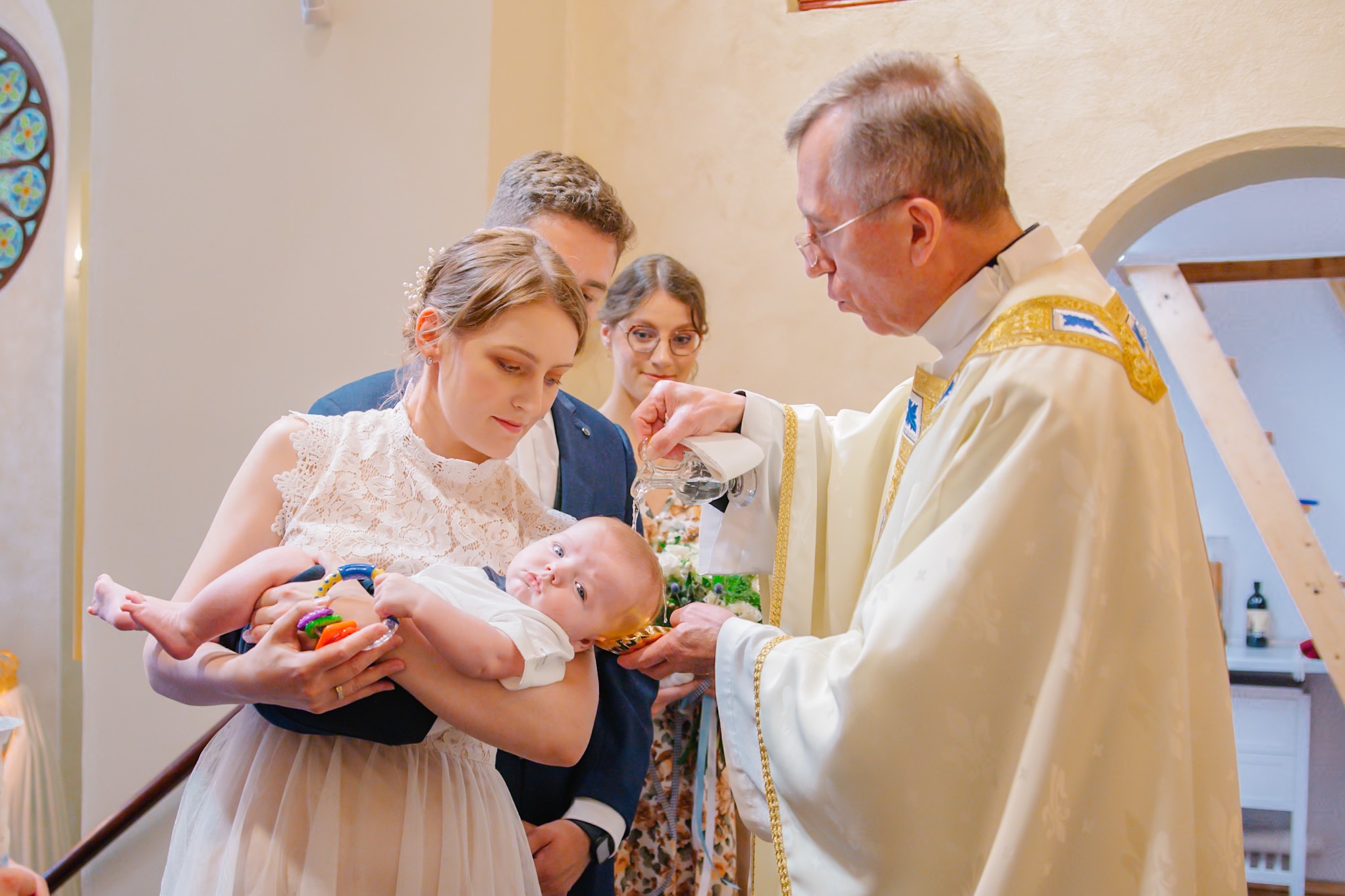 Chrzest dziecka w kościele: matka trzyma niemowlę, ksiądz polewa wodą z naczynia, w tle rodzina. Uroczystość w jasnych barwach, delikatne oświetlenie.