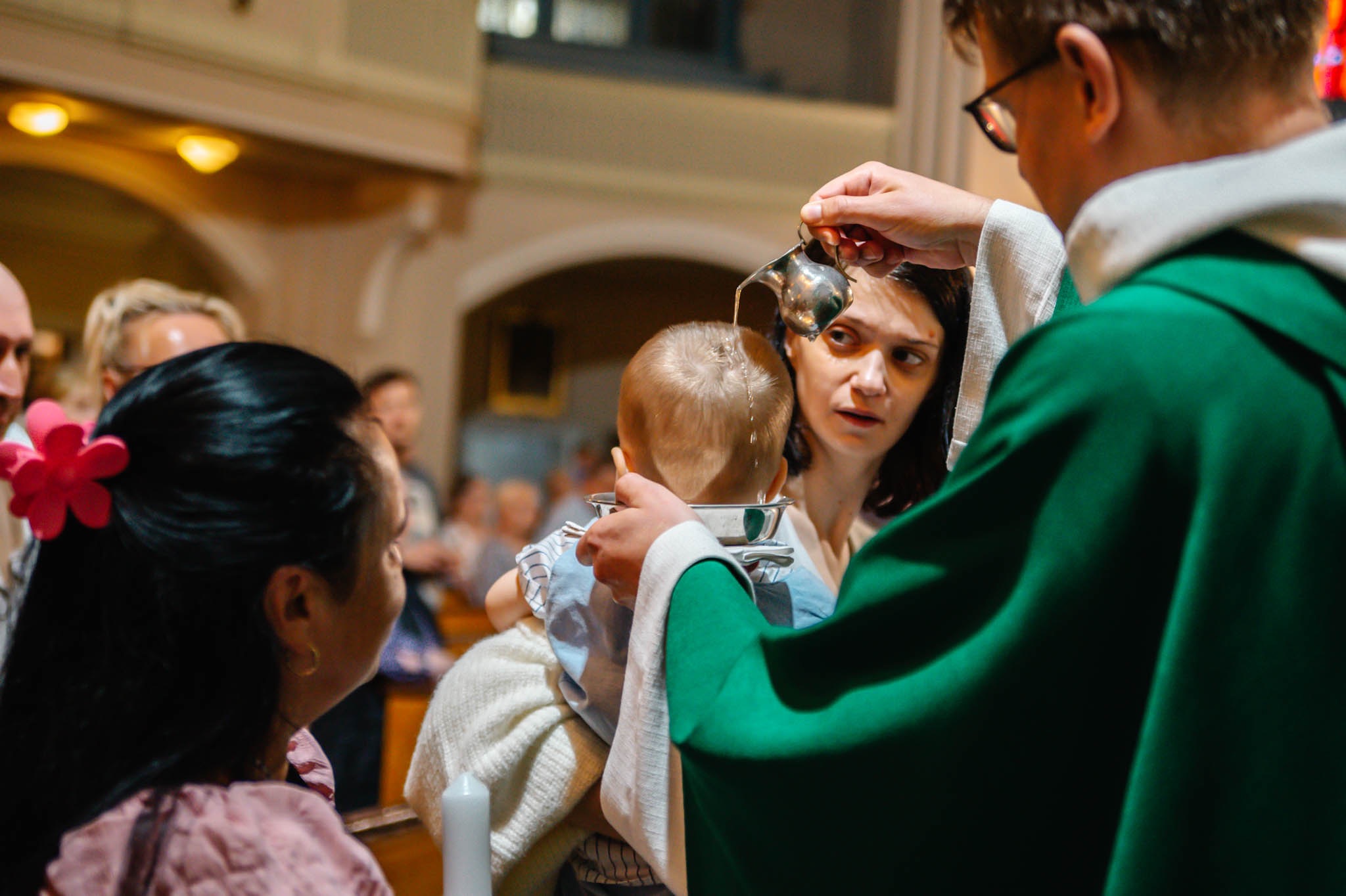 Chrzest dziecka w kościele: ksiądz polewa wodą główkę niemowlęcia, matka z uwagą obserwuje. W tle widoczni goście i detale architektoniczne wnętrza kościoła.