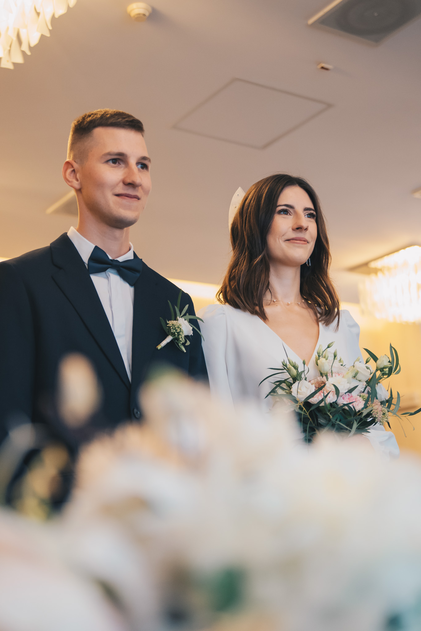 Elegancka para młoda podczas ceremonii: pan młody w granatowym smokingu, panna młoda w białej sukni z bukietem. Delikatne światło, subtelny uśmiech.