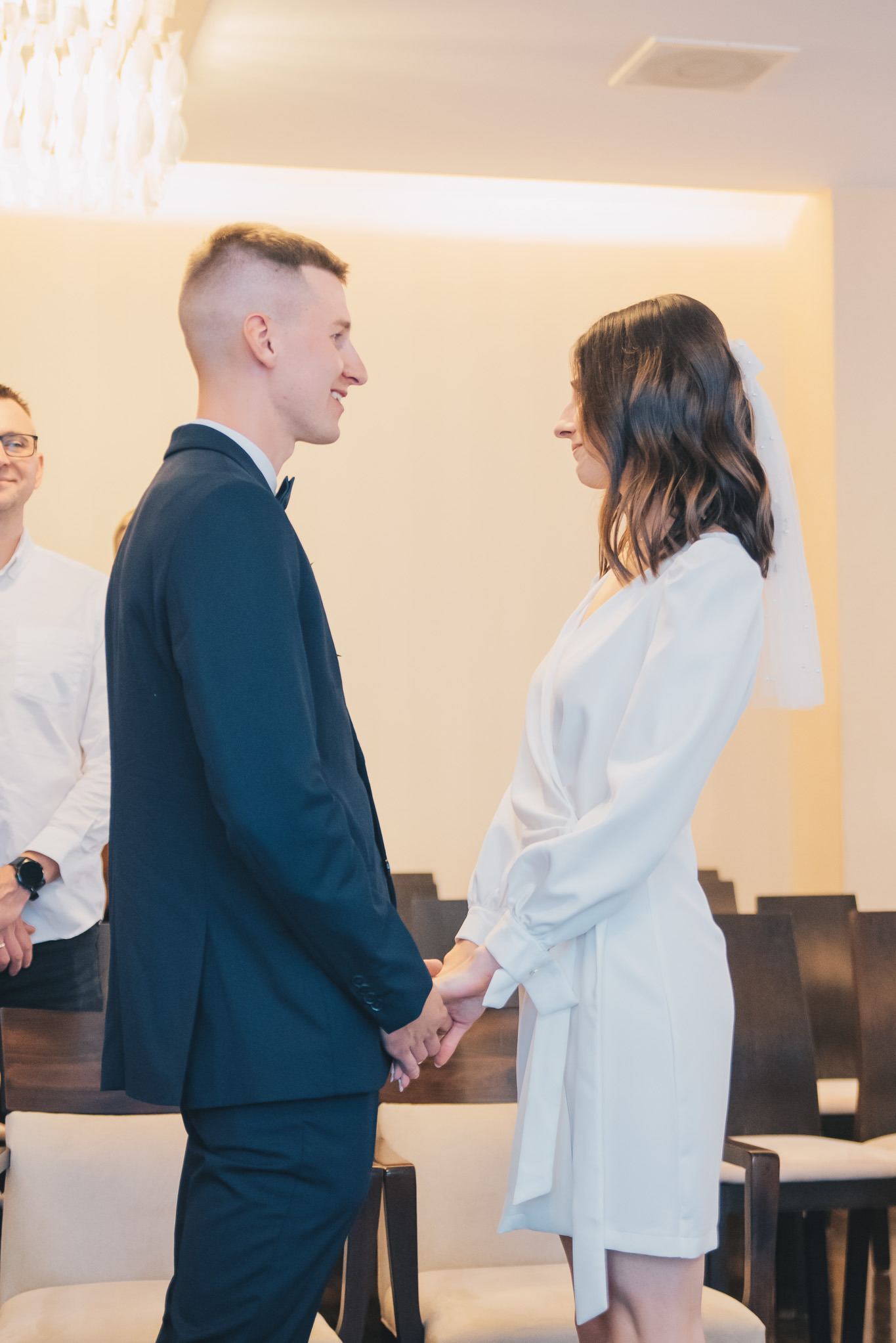 Młoda para trzymająca się za ręce podczas ceremonii ślubnej w jasnym wnętrzu. Panna młoda w białej sukience, pan młody w granatowym garniturze, profilowe ujęcie.