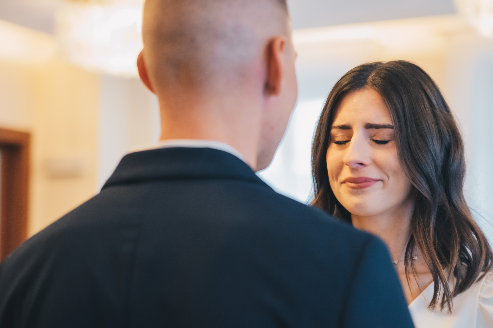 Panna młoda w białej sukni, z zamkniętymi oczami i wzruszeniem, podczas ceremonii ślubnej. Widoczny fragment pleców pana młodego w ciemnym garniturze.