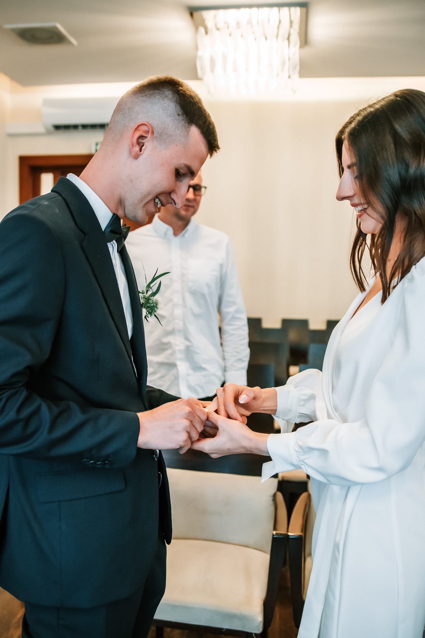 Para młoda wymienia się obrączkami podczas ceremonii ślubnej w jasnym wnętrzu, z uśmiechem i świadkiem w tle. Elegancki garnitur pana młodego i biała suknia panny młodej.