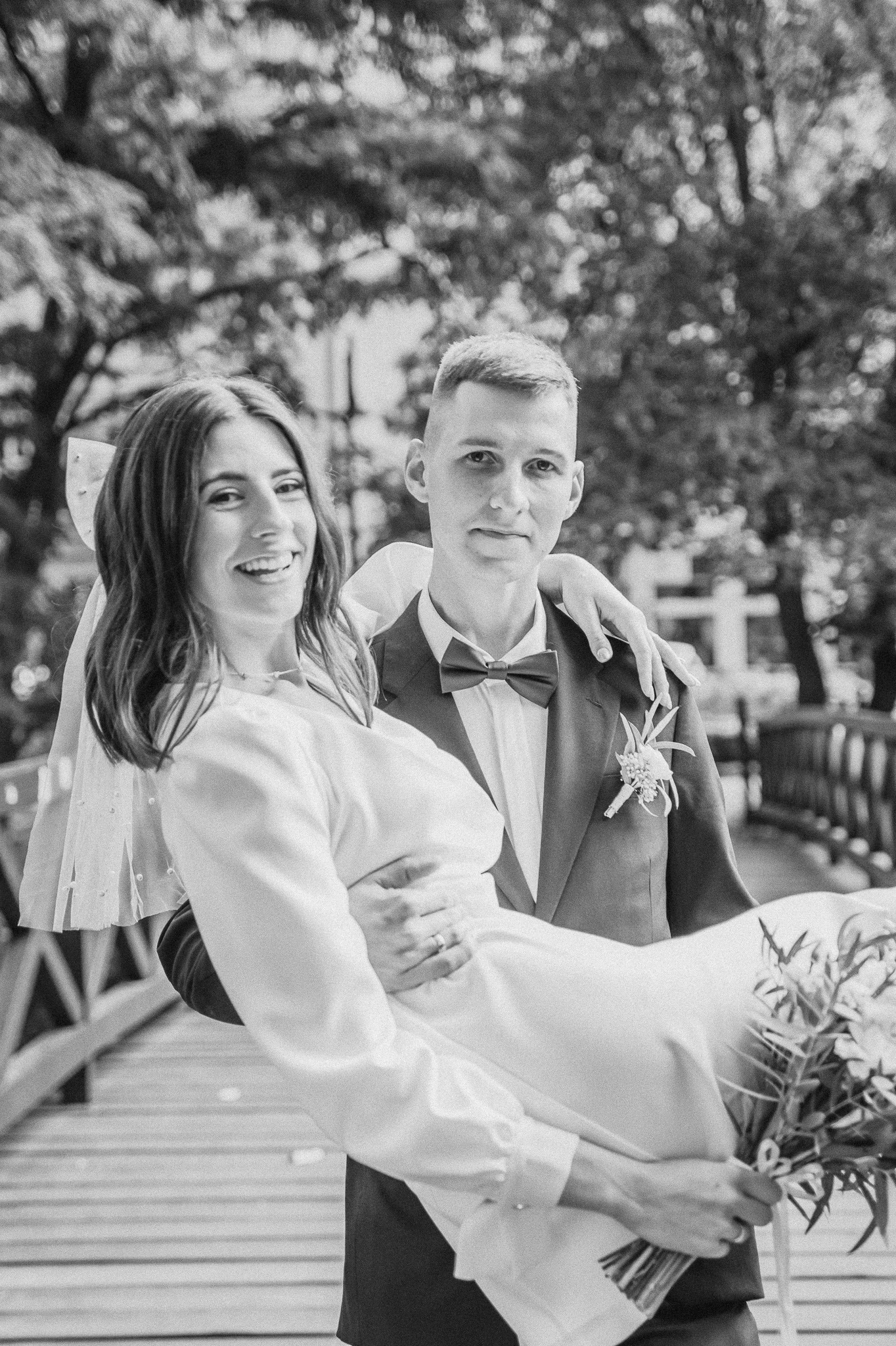 Czarno-białe zdjęcie pana młodego trzymającego uśmiechniętą pannę młodą na rękach, z bukietem i welonem, na drewnianym moście w plenerze.